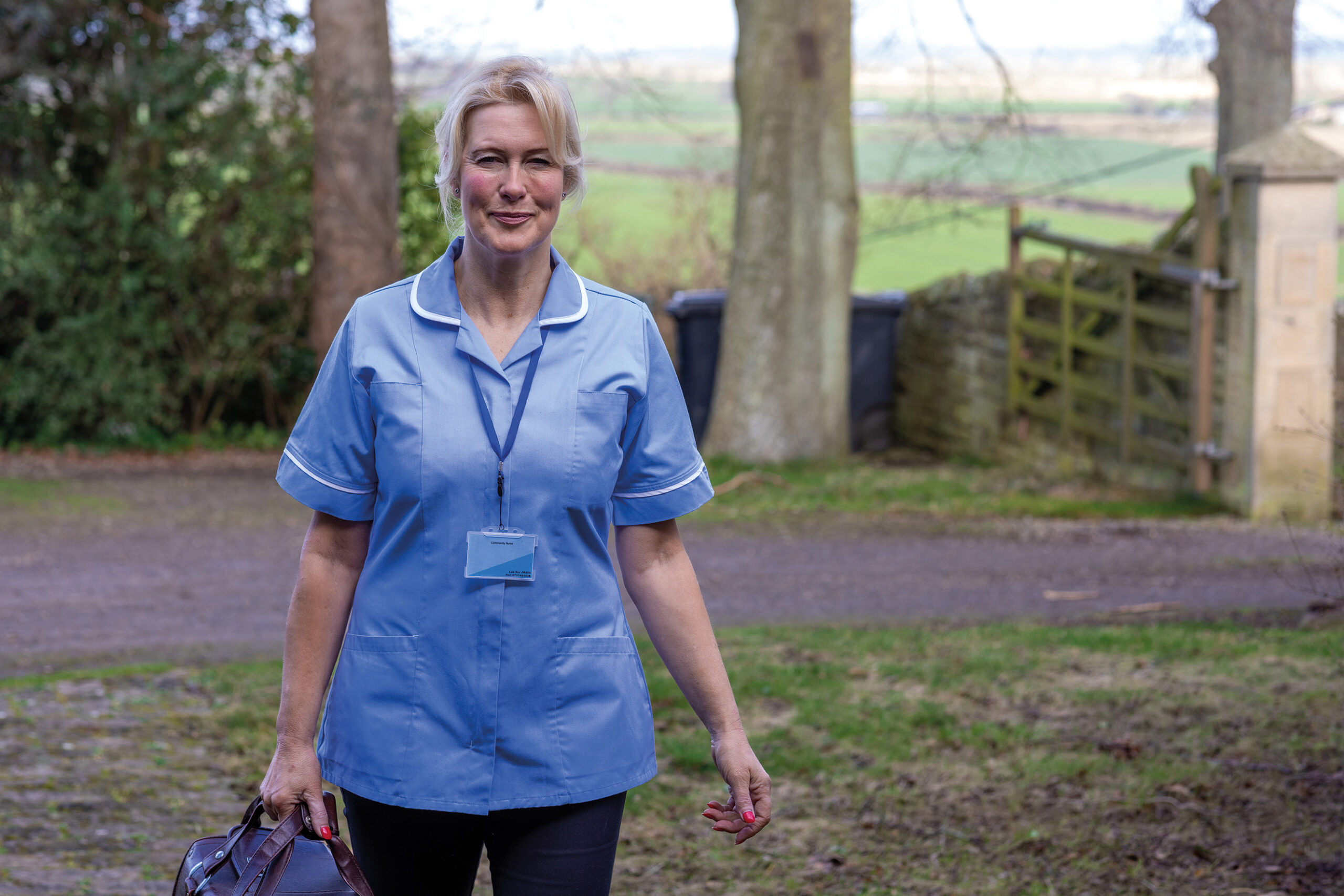 Three-quarter length portrait of a healthcare worker standing looking into the camera. She is smiling in her uniform with a rural scene behind her. She is in the Northeast of England.